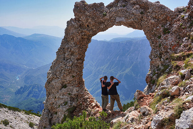Zwei junge Männer haken sich unter und blicken aus einem Torbogen auf die umliegenden Berge im Blidinje-Nationalpark in Bosnien-Herzegowina