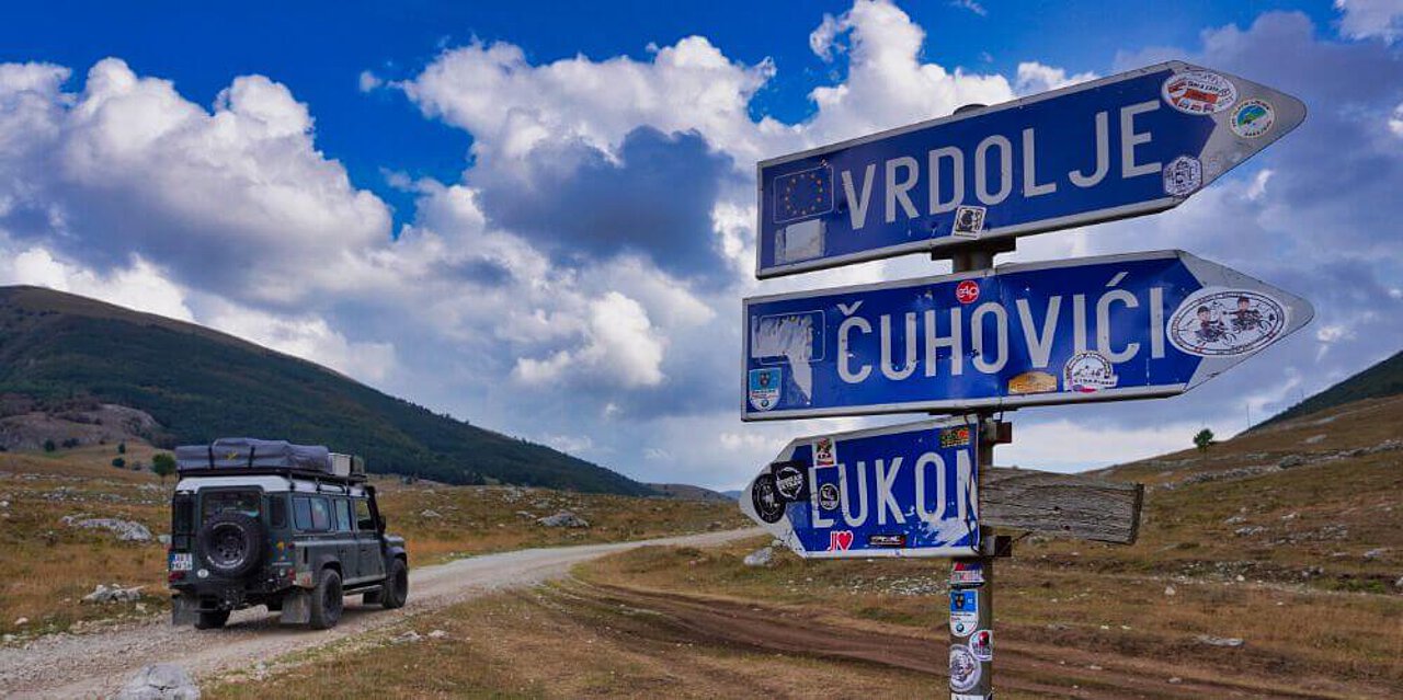 Ein blaues Schild weist den Weg zu den Bergdörfern von Bosnien-Herzegowina, im Hintergrund steht ein Landrover